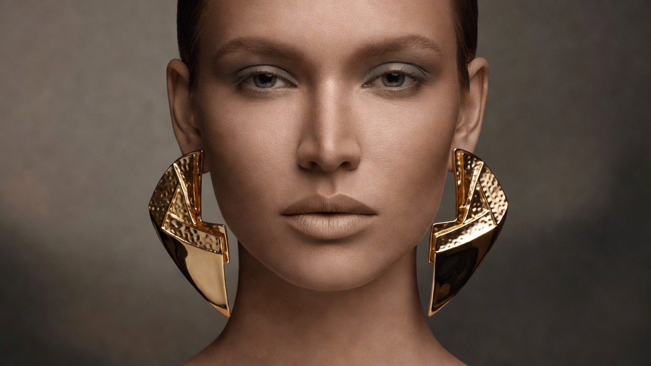 Portrait of a person wearing large sculptural gold earrings with softly defined eyes and a neutral lip